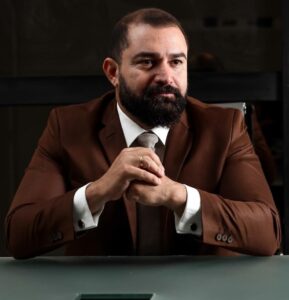 Advogado criminalista de postura confiante e foco estratégico, usando terno marrom e gravata, sentado em ambiente corporativo, transmitindo profissionalismo e autoridade jurídica. Ideal para sites e blogs de advocacia penal, defesa criminal, branding pessoal e marketing jurídico.