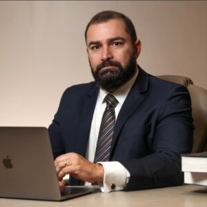 Advogado experiente em escritório jurídico, vestido com terno escuro e gravata, trabalhando em notebook Apple sobre mesa com livros jurídicos. Imagem transmite profissionalismo, seriedade e autoridade no Direito. Ideal para sites de advocacia, páginas institucionais e marketing jurídico voltado para advocacia penal, patrimonial ou empresarial.